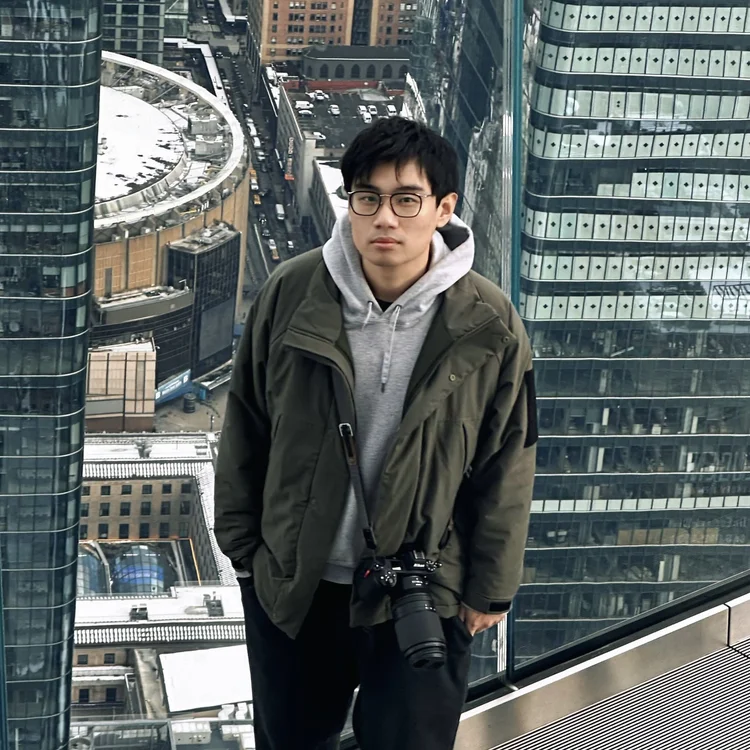 Yipeng Wang wearing a gray hoodie and green jacket, standing in a tall building with a camera hanging from his neck, overlooking a cityscape.